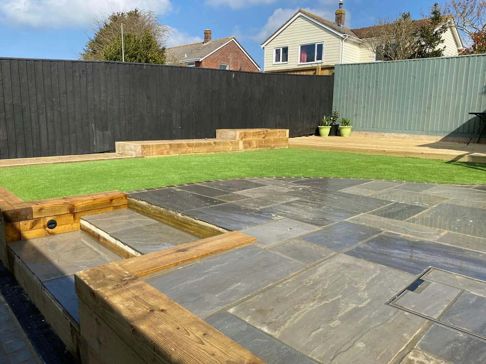 A grey stone patio with timber sleeper edging, below an artificial lawn