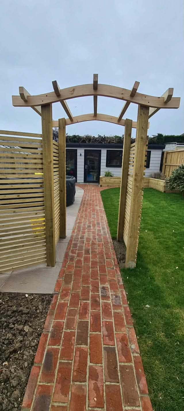 A brick path running under a timber pergola, between slatted screens and lawn