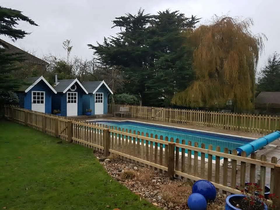 Timber picket fencing around an outdoor swimming pool, with blue beach-hut style changing rooms behind