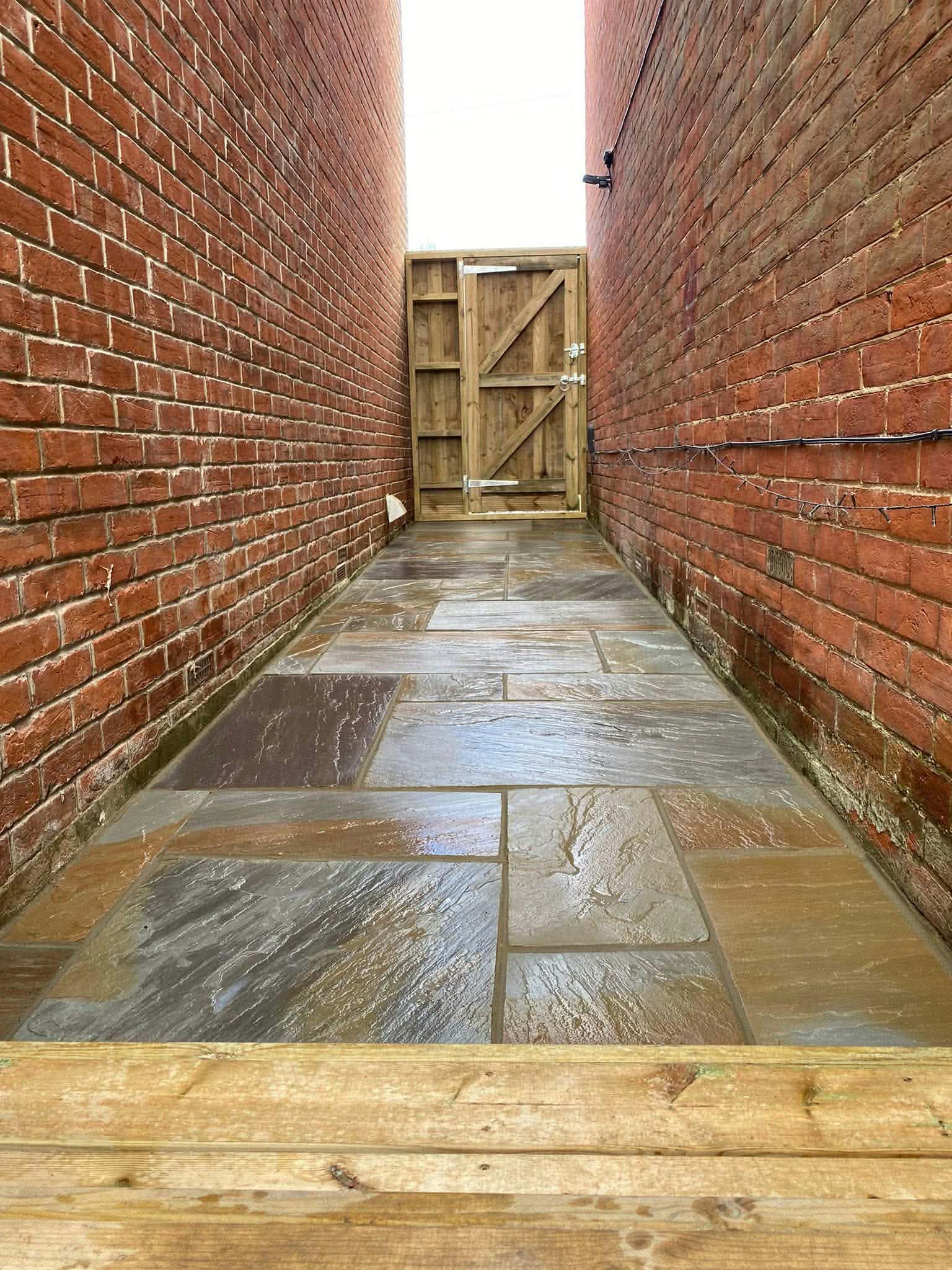 A narrow sandstone-paved side path between two brick walls, with a new timber gate at the end